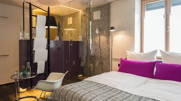 A modern hotel room with a large bed, a glass shower enclosure, a small table with a chair, and a large window.
