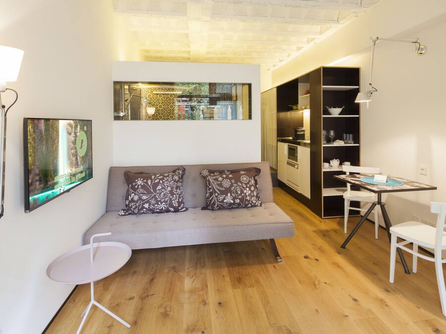 A modern living room with a grey sofa, two patterned cushions, a small white table, and a dining area with a white table and chairs.