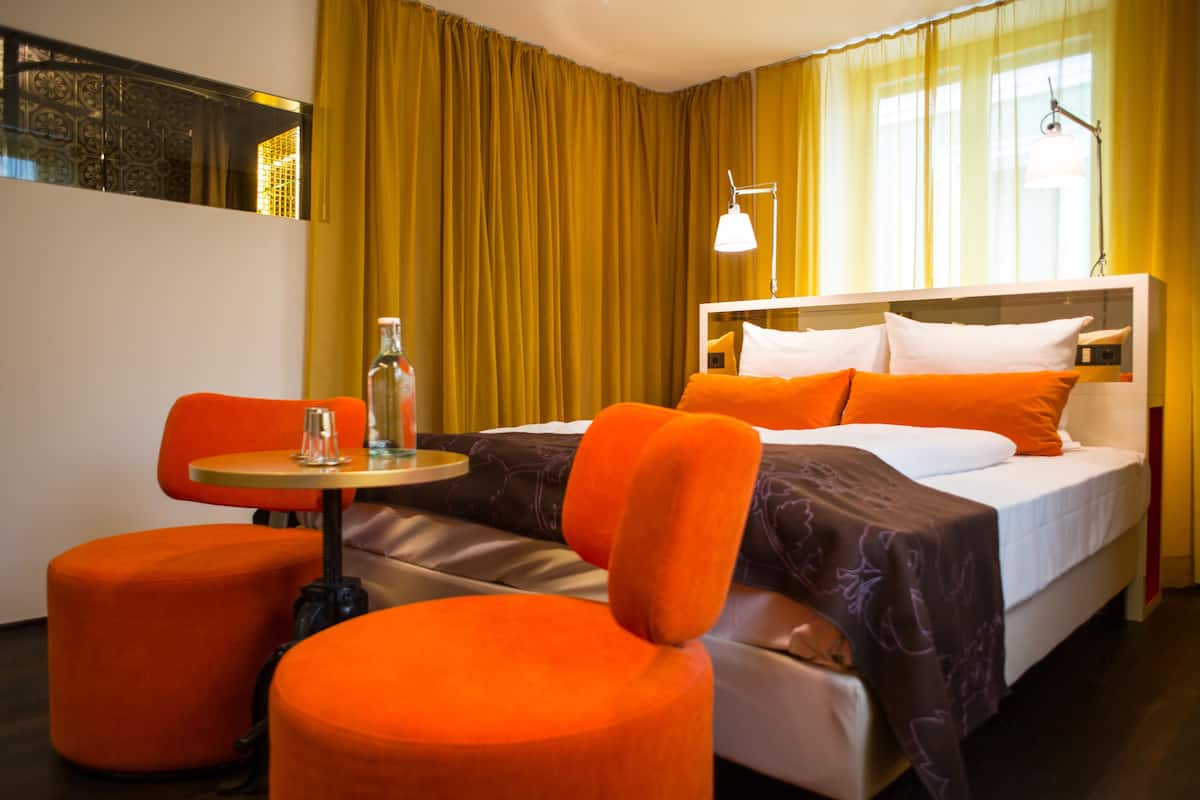 A hotel room with a bed, orange chairs, a small table, and a bottle.