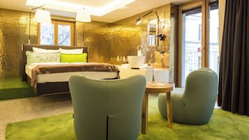 A modern hotel room with a large bed, two armchairs, a small table, and a bathroom with a sink and mirror.