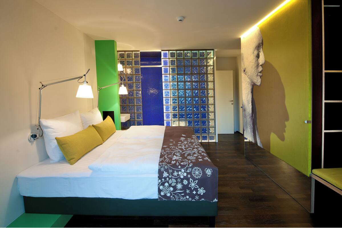 A modern hotel room with a neatly made bed, a bedside lamp, a green accent wall, and a view of a blue patterned wall.