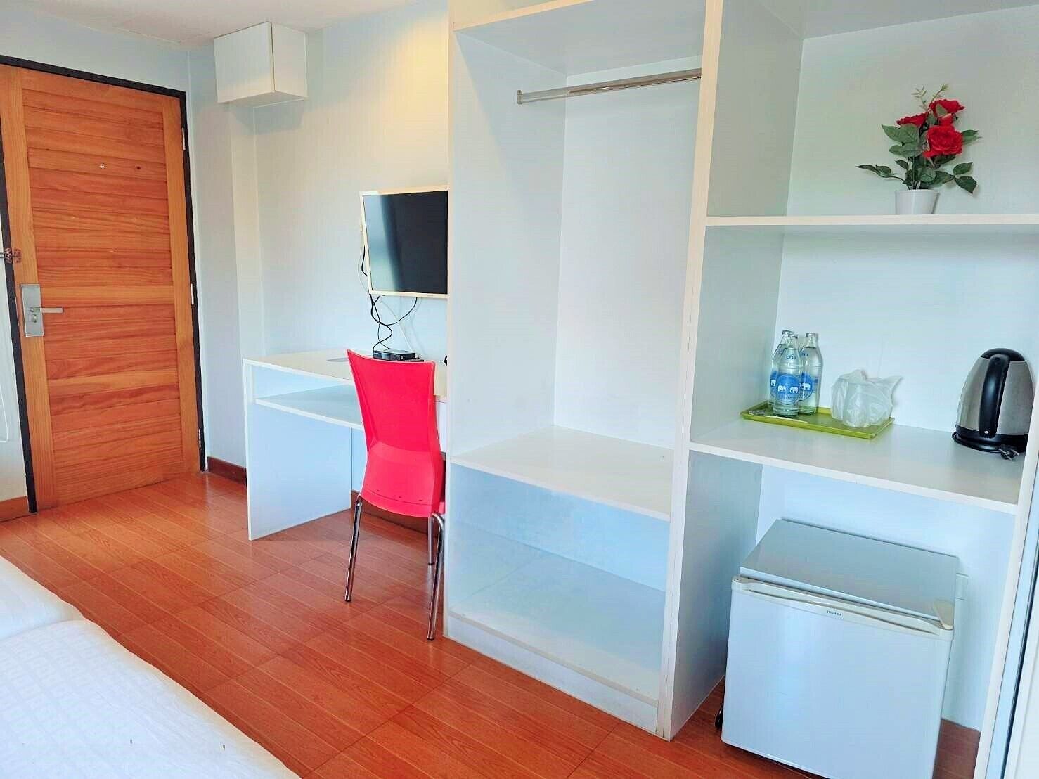 A compact room with a wooden door, a red chair, a small white refrigerator, a shelf with a vase of flowers, and a kettle.