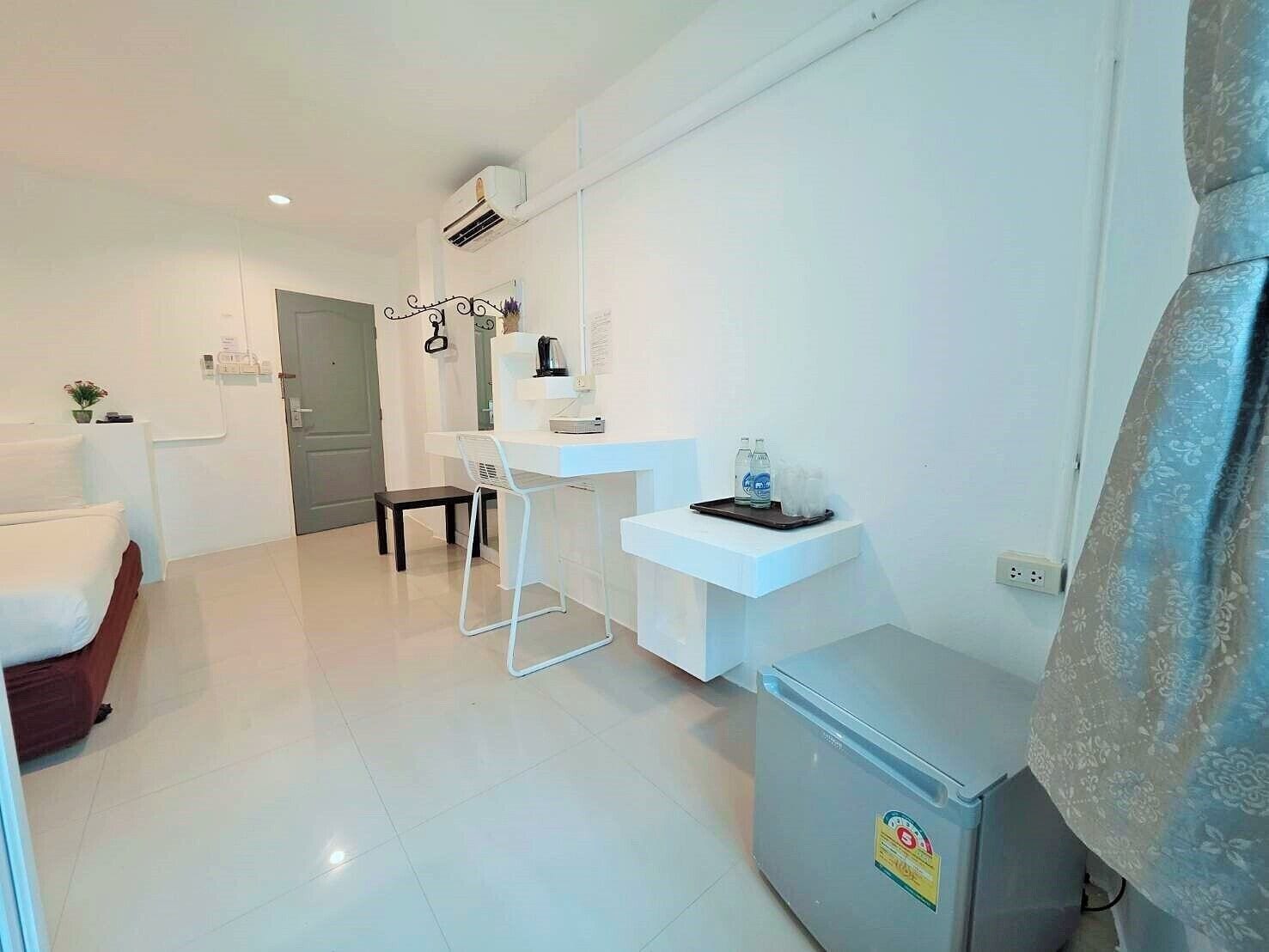 Una habitación moderna y luminosa con una cama, una mesita con una silla, un refrigerador y un estante en la pared con una tetera y tazas.