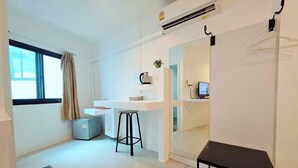 Een moderne, goed verlichte kamer met een bureau, een computer en een klein krukje. Er is een raam met gordijnen, een radiator en een wandairconditioner.