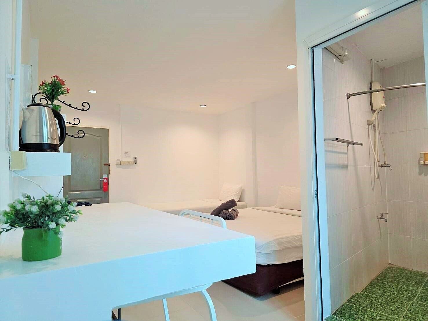 A hotel room with a white bed, a small table with a plant, a shower area, and a white wall.
