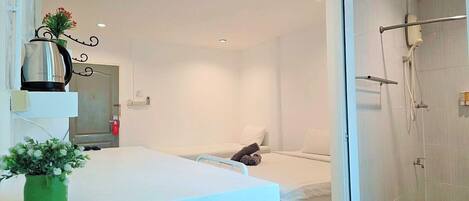 A hotel room with a white bed, a small table with a plant, a shower area, and a white wall.