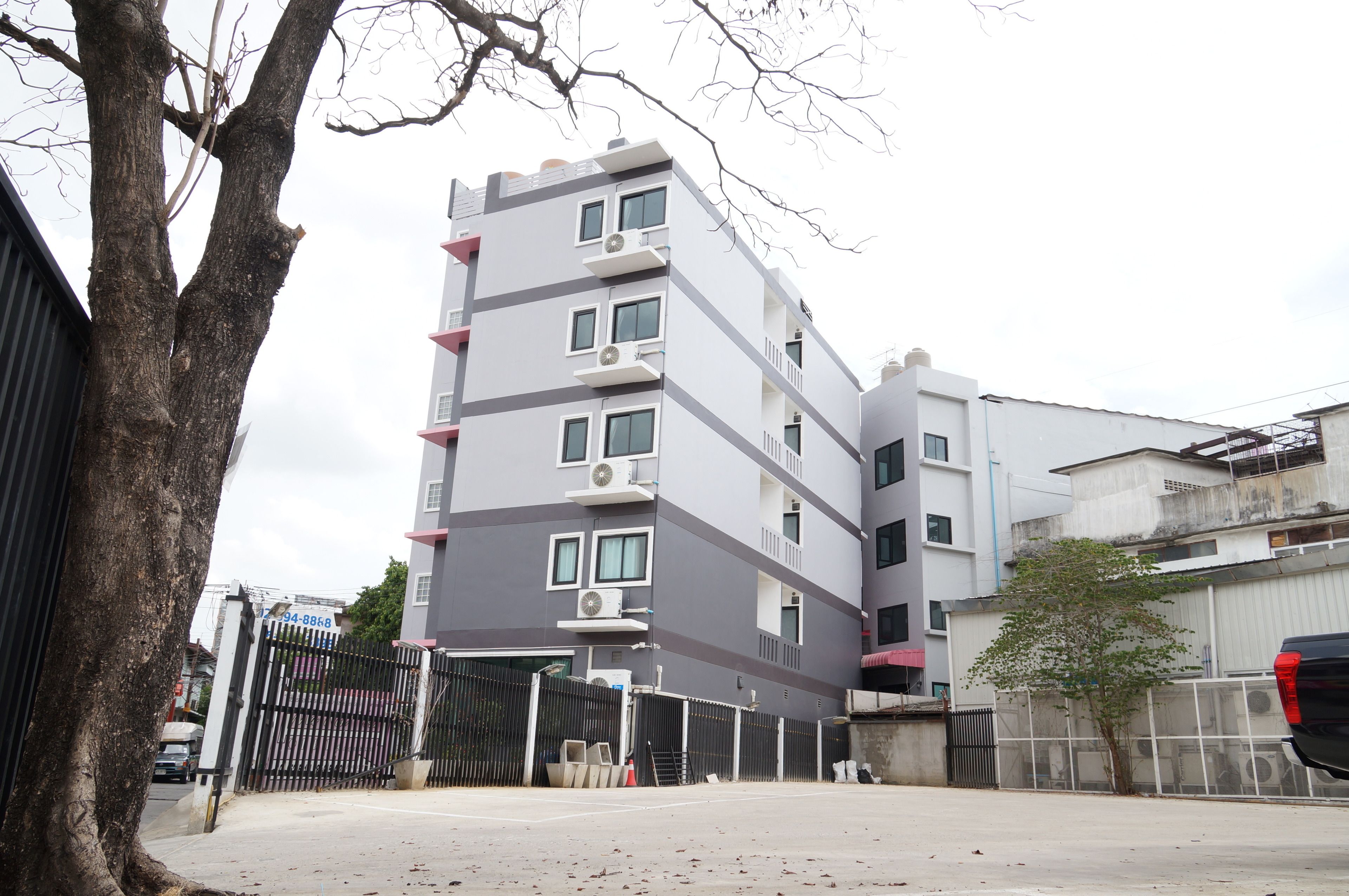 A multi-story building with a gated entrance, a tree in the foreground, and a parked vehicle.