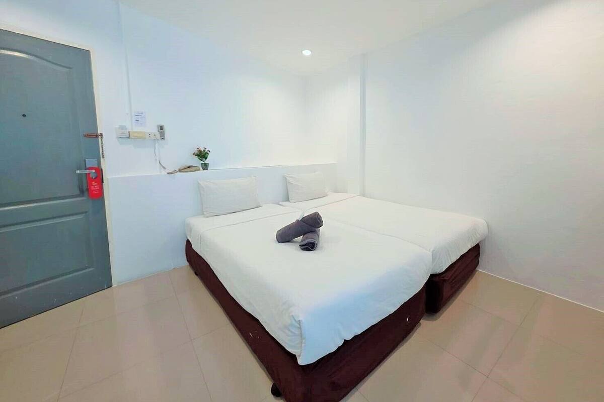 Une chambre d’hôtel avec un lit simple, une table de chevet, une petite plante en pot et une porte avec une poignée rouge.