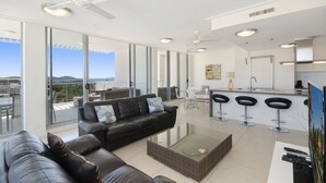 Un salon moderne avec un canapé en cuir, une table basse en verre et un coin cuisine comprenant un bar et des tabourets hauts.