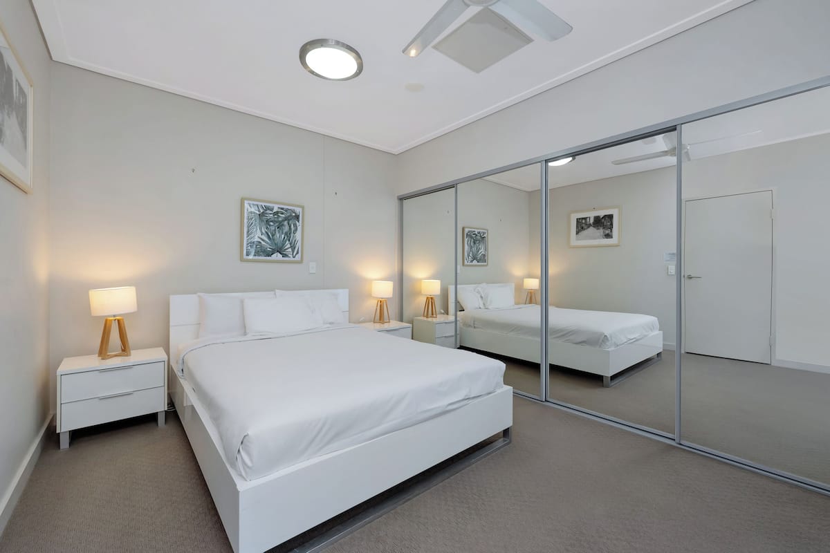 A modern hotel room with a large bed, bedside tables with lamps, a wardrobe, and a large mirror.
