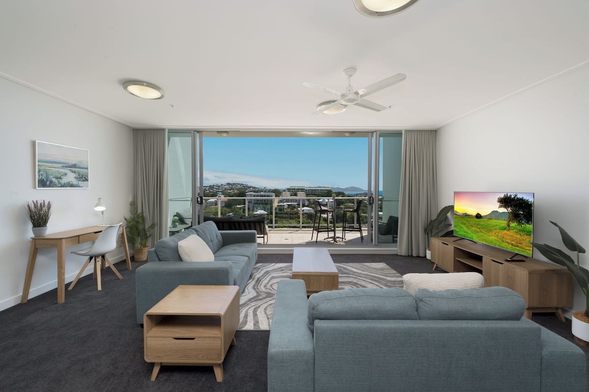 A modern living room with a grey sofa set, a wooden coffee table, and a TV on a wooden stand. There is a balcony with a view of a cityscape.