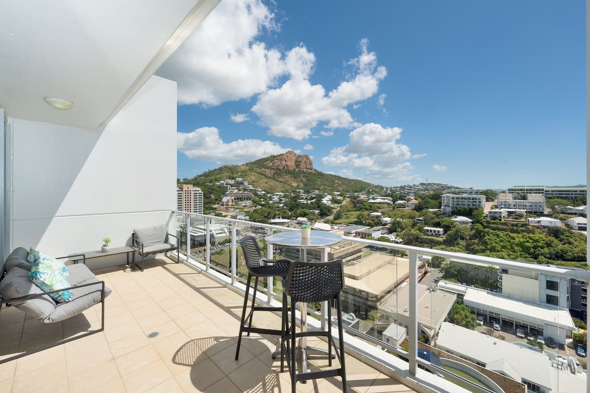 A balcony with a table and chairs, overlooking a cityscape with buildings and a mountain.