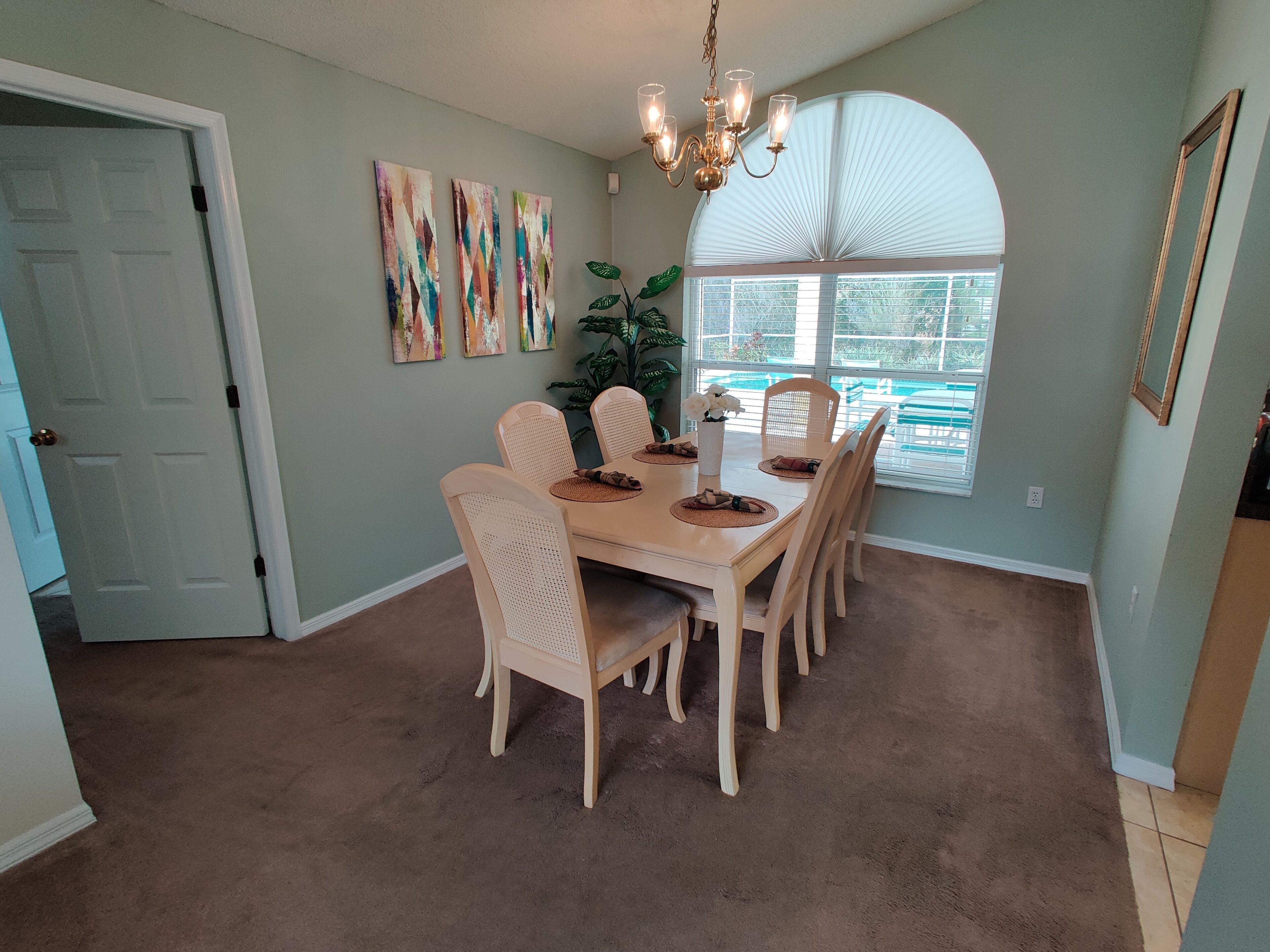 Un coin repas avec un tapis beige, une table en bois et six chaises. Trois tableaux abstraits ornent le mur, ainsi qu’un lustre et une fenêtre équipée de stores.