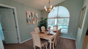A dining area with a beige carpet, a wooden table, and six chairs. There are three abstract paintings on the wall, a chandelier, and a window with blinds.
