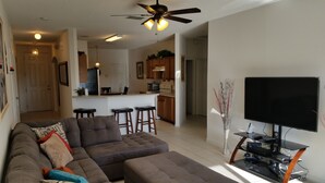 A living room with a sectional sofa, ottoman, and a flat-screen TV on a stand. There is a ceiling fan, a kitchen in the background, and a hallway leading to a door.