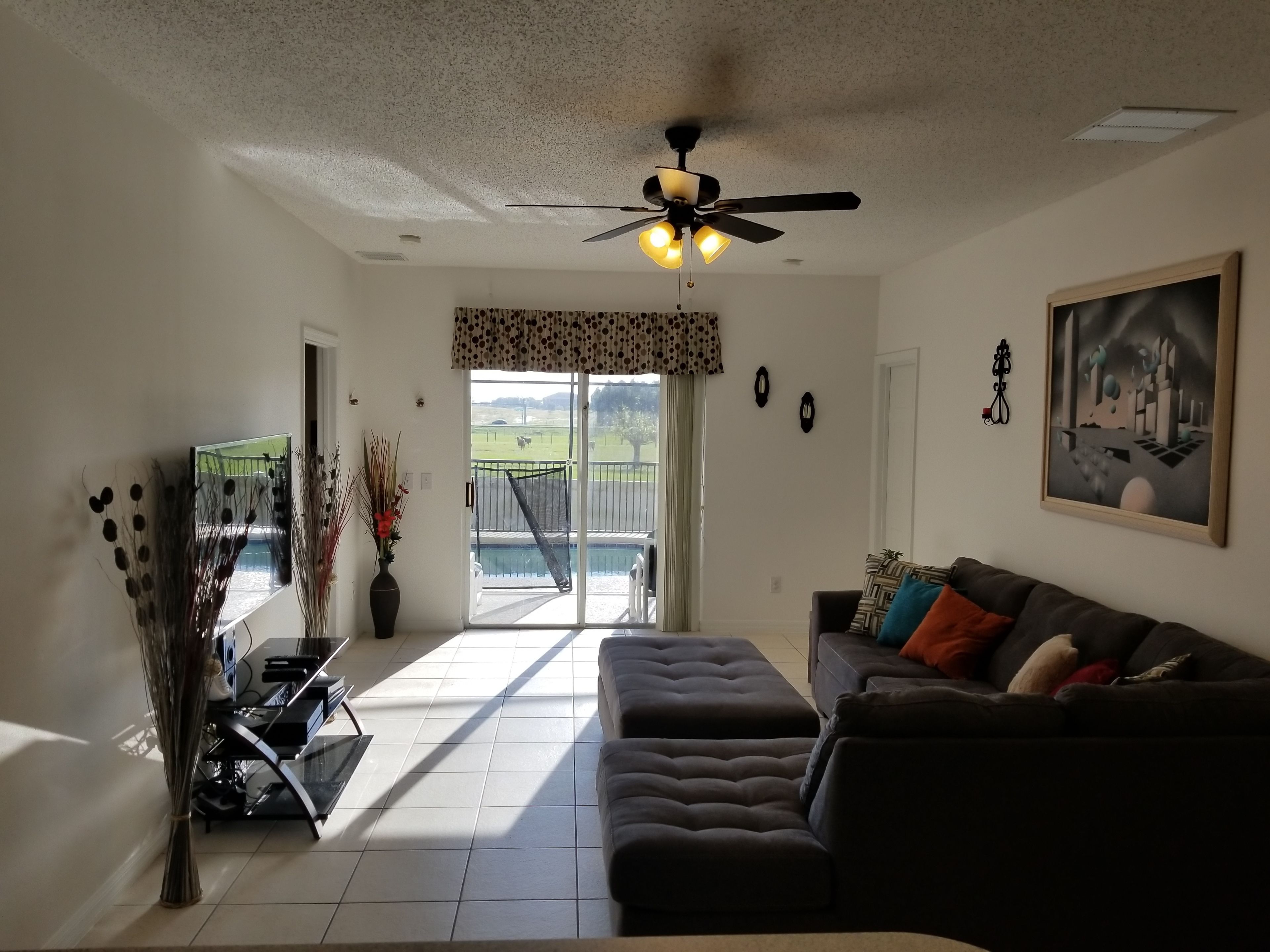 A living room with a dark-colored sofa, a ceiling fan, a TV, and a view of a pool and outdoor area.