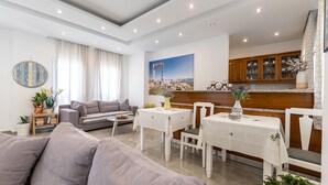 A modern living room with a sofa, a dining table with chairs, a kitchen area with wooden cabinets, and a large painting on the wall. - Sun Beach Hotel (Naxos)