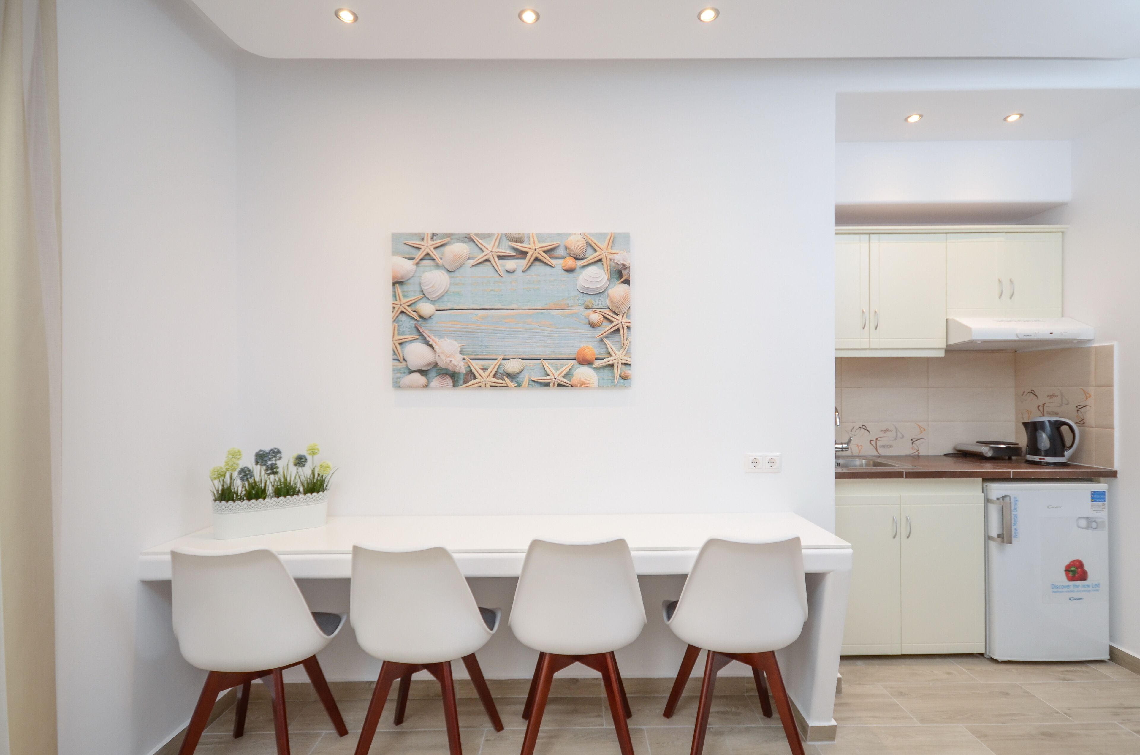 Una cocina moderna con armarios blancos, encimera blanca y un refrigerador pequeño. Hay una mesa de comedor con cuatro sillas blancas y un elemento decorativo en la pared sobre ella.