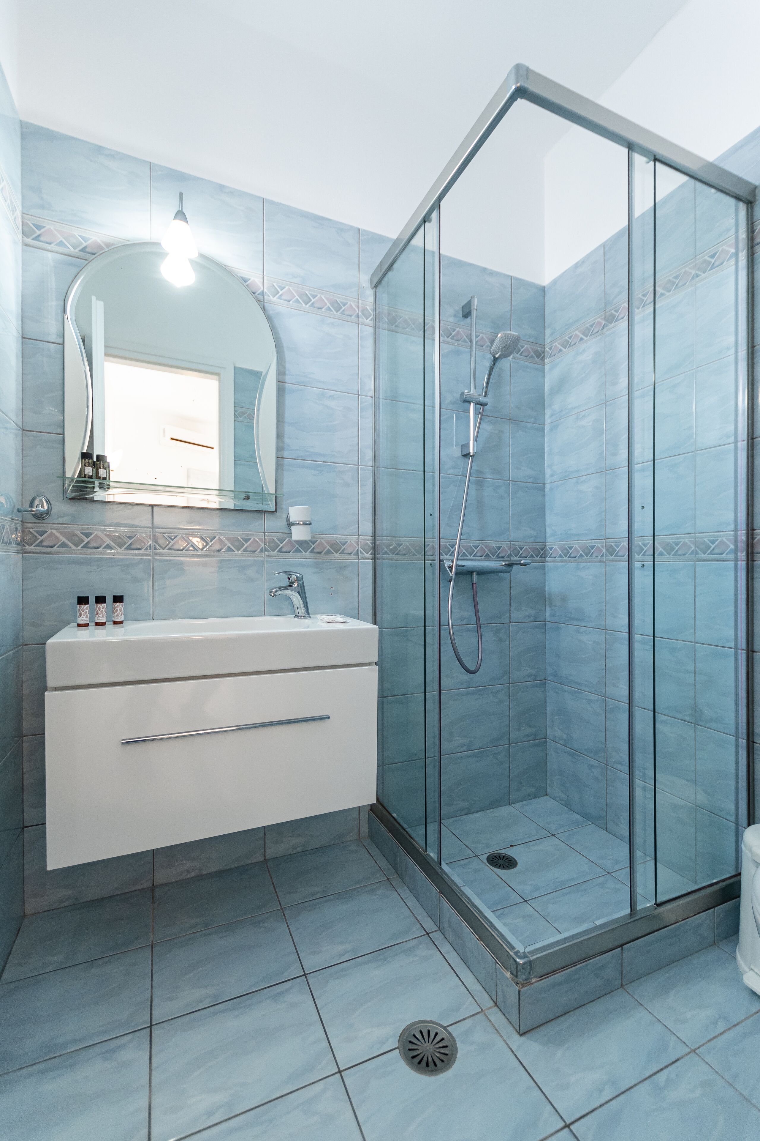 Un baño con ducha envuelta en vidrio, un lavamanos blanco con un espejo encima y paredes de azulejos azules.