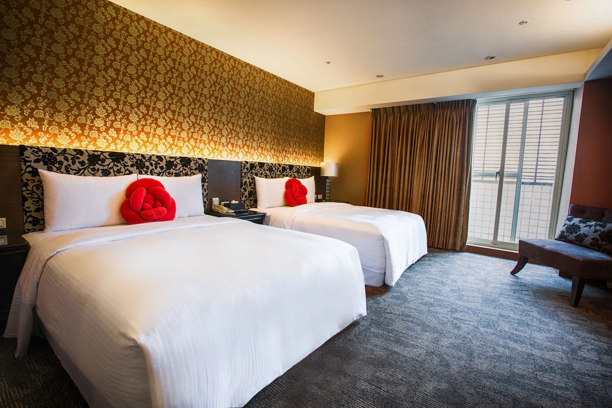 A hotel room with two beds, a large window with curtains, a desk with a lamp, and a chair.
