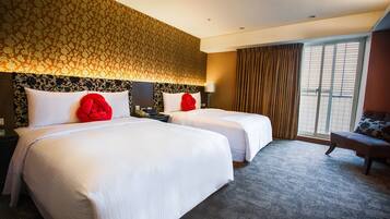 Quarto de hotel com duas camas, uma janela grande com cortinas, uma mesa com abajur e uma cadeira.