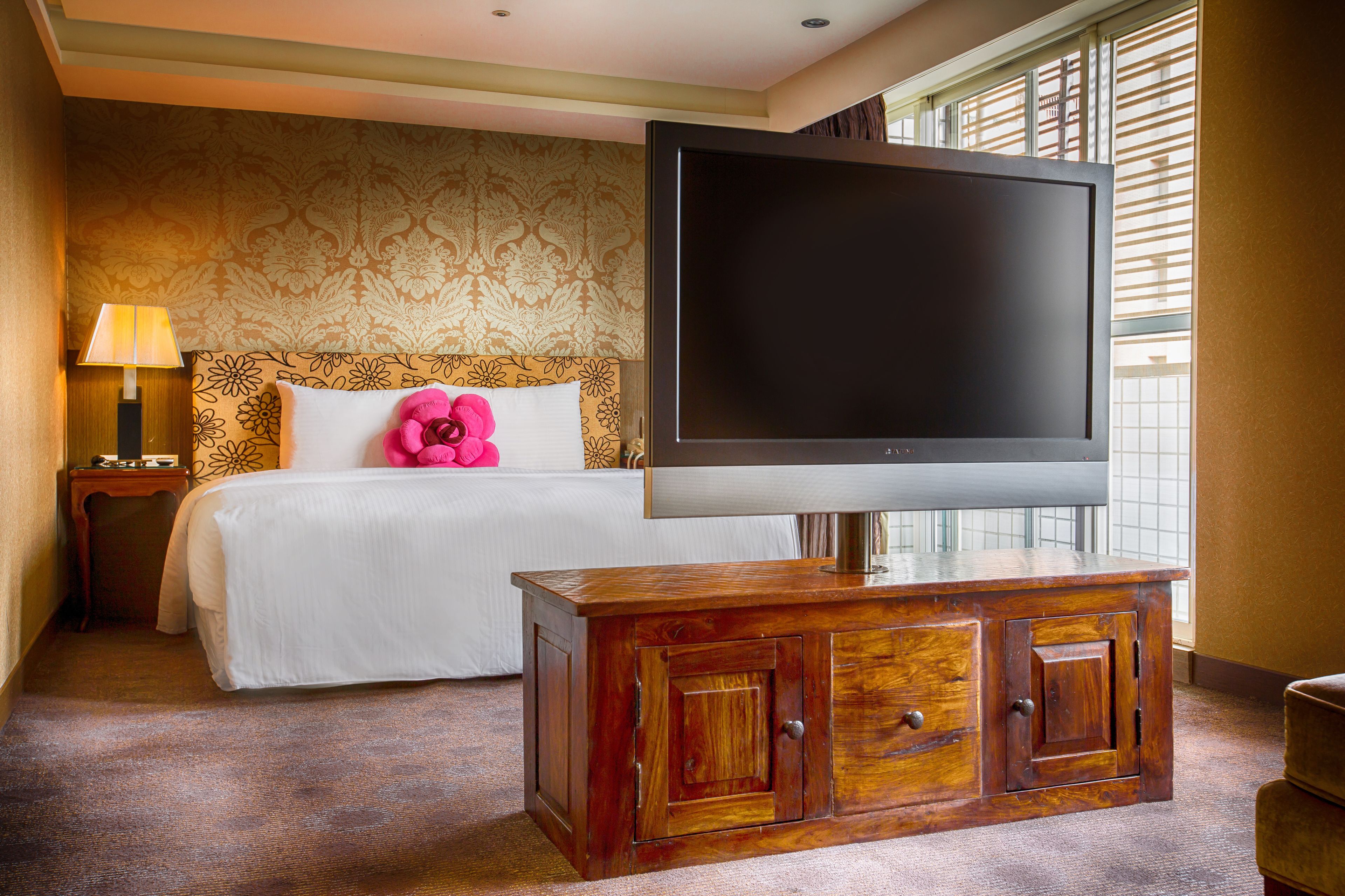 Quarto de hotel com cama, televisão sobre um armário de madeira, mesa de cabeceira com abajur e janela com persianas.