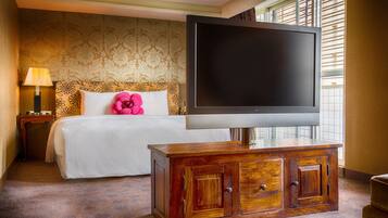 Quarto de hotel com cama, televisão sobre um armário de madeira, mesa de cabeceira com abajur e janela com persianas.