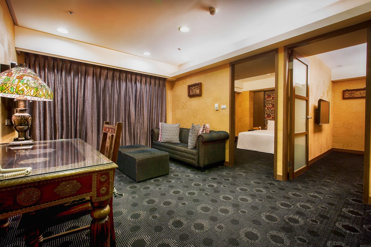 A hotel room with a glass-top table, a sofa with patterned cushions, a patterned carpet, and a door leading to a bathroom.