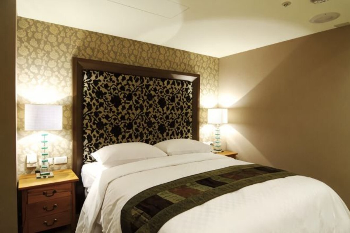 A hotel room with a patterned headboard, a bed with white linens, two bedside tables with lamps, and a nightstand with drawers.