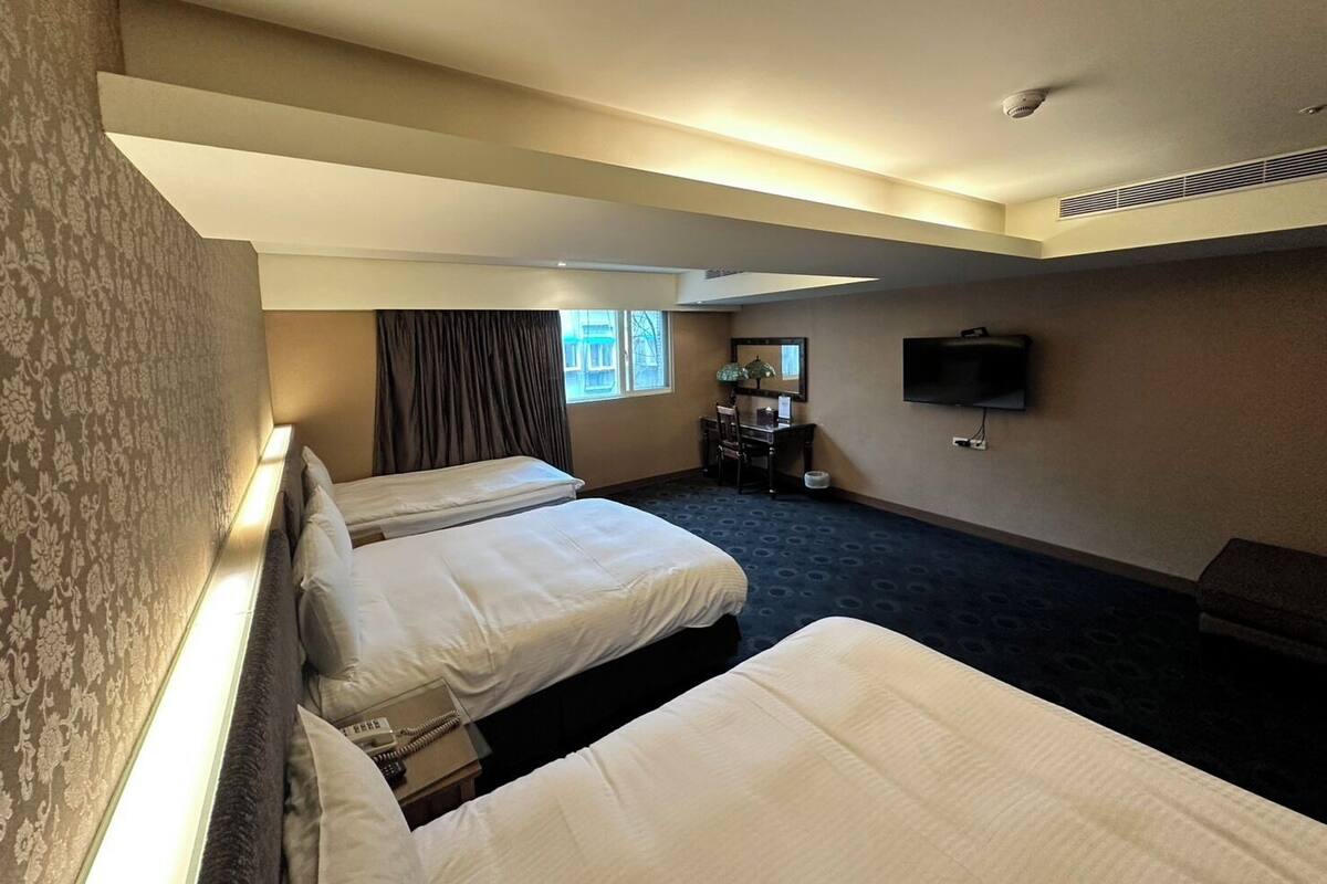A hotel room with three beds, a desk with a chair, a television, and a window with curtains.