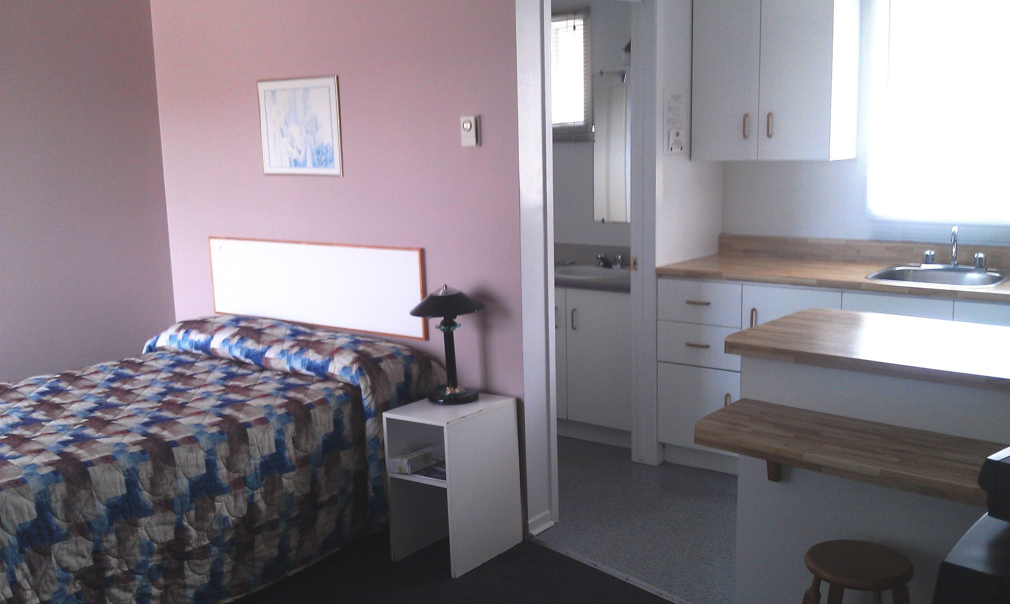 Un dormitorio con cama, mesita de noche y un pequeño escritorio con lámpara. Cuenta con una kitchenette con lavabo y armarios.