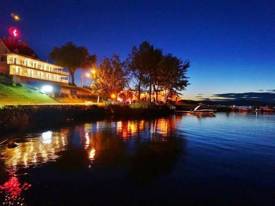 A waterfront property with a large house, trees, and a calm body of water reflecting lights.