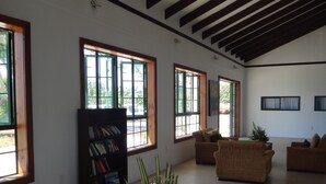 A spacious living area with large windows, a bookshelf, wicker furniture, and a central table with a potted plant. - Amoa Resort Savaii (Tuasivi)
