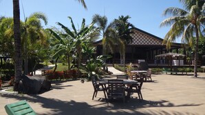 An outdoor seating area with tables and chairs, surrounded by palm trees and a building in the background. - Amoa Resort Savaii (Tuasivi)