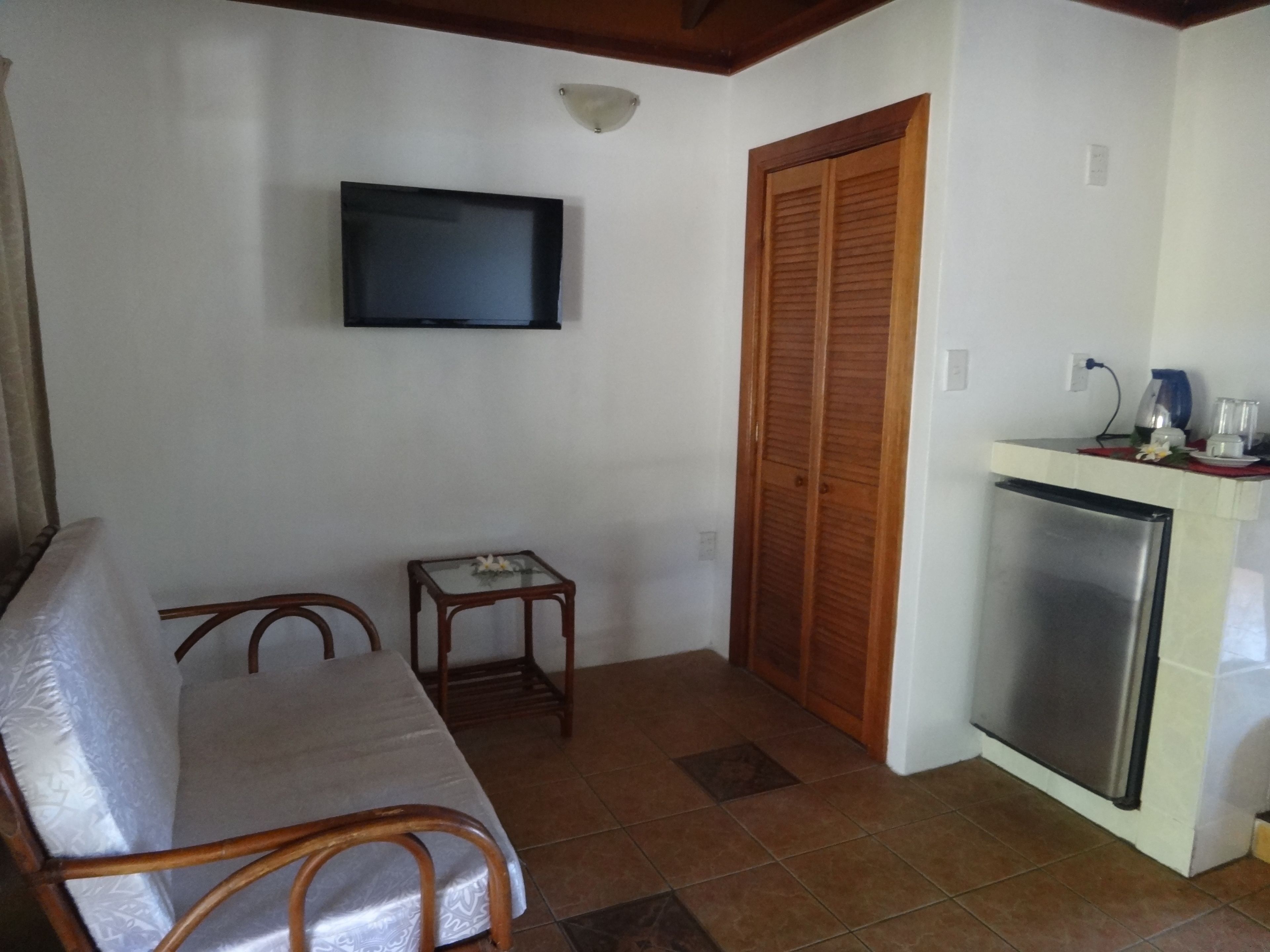 A room with a sofa, a small table, a television mounted on the wall, a wooden door, and a refrigerator.