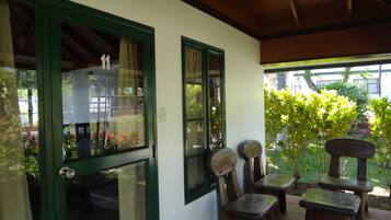 Un patio cubierto con una puerta verde, dos ventanas y vistas a un jardín. Se observan tres taburetes de madera y un banco.