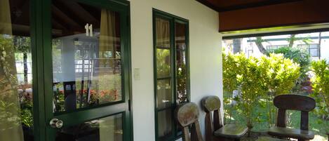 A covered patio area with a green door, two windows, and a view of a garden. There are three wooden stools and a bench.