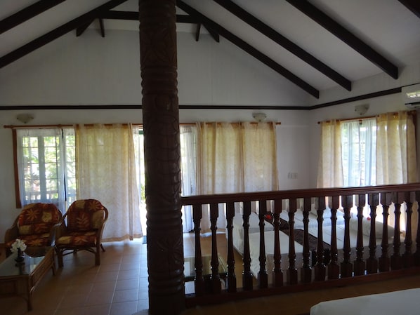 Una habitación amplia con techo alto, vigas de madera, una escalera con barandilla de madera y grandes ventanales con cortinas translúcidas.