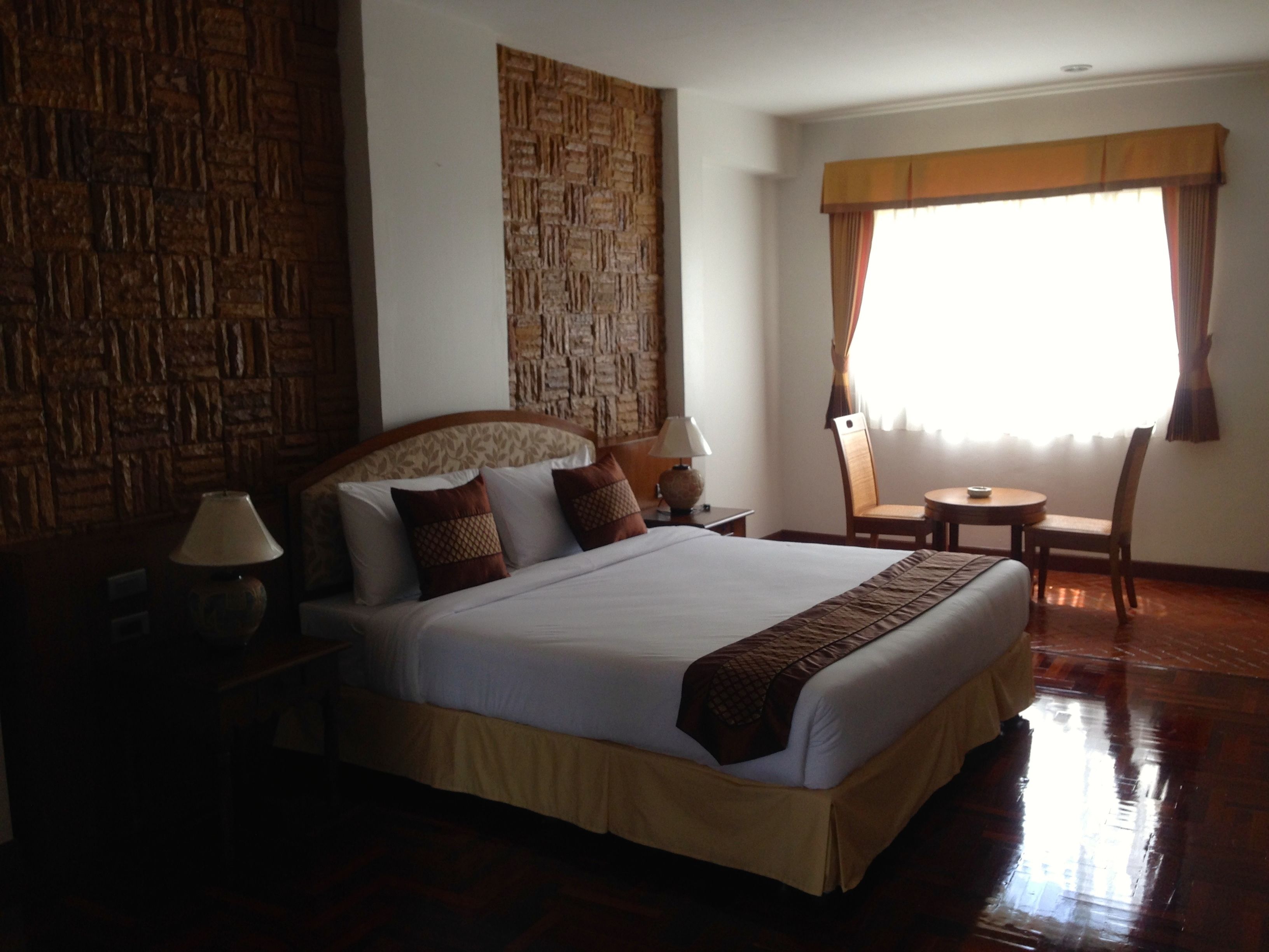 A hotel room with a large bed, a wooden headboard, a small table with a chair, and a window with curtains.