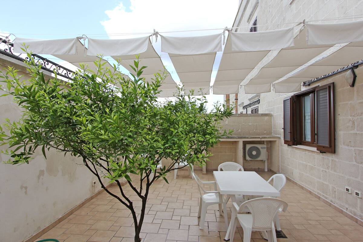 An outdoor patio area with a white table and chairs, a small tree, and a white awning providing shade.