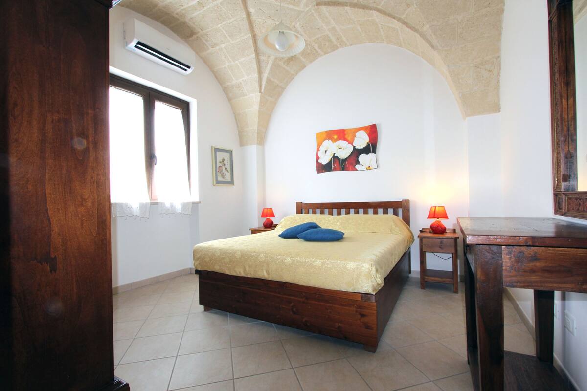 A bedroom with a wooden bed, two bedside lamps, a painting on the wall, and a tiled floor.