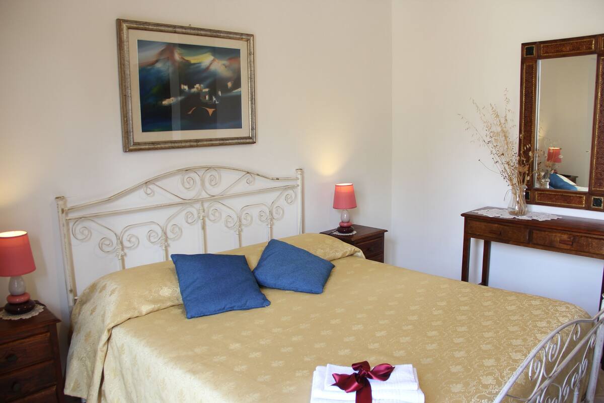 A neatly made bed with a patterned bedspread, two pillows, a bedside lamp, a framed painting on the wall, and a mirror above a dresser.