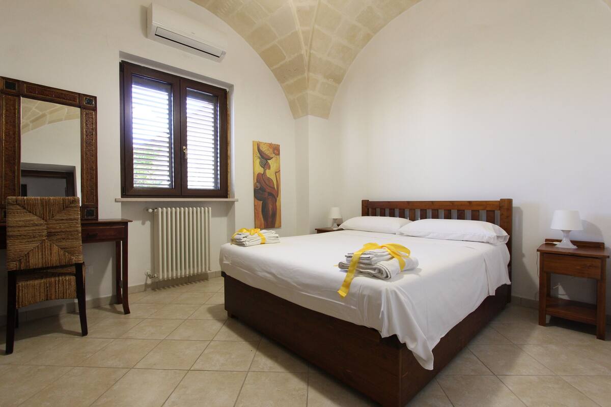 A bedroom with a large bed, a wooden chair, a desk, a window with blinds, a radiator, and a painting on the wall.