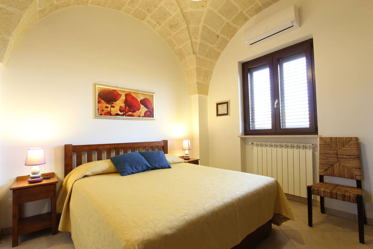 A bedroom with a large bed, two bedside lamps, a framed picture on the wall, a window with blinds, and a radiator.