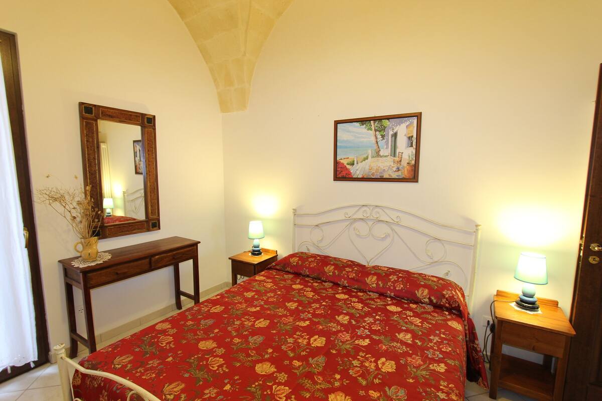 A bedroom with a bed, a bedside table with a lamp, a dresser, a mirror, and a painting on the wall.