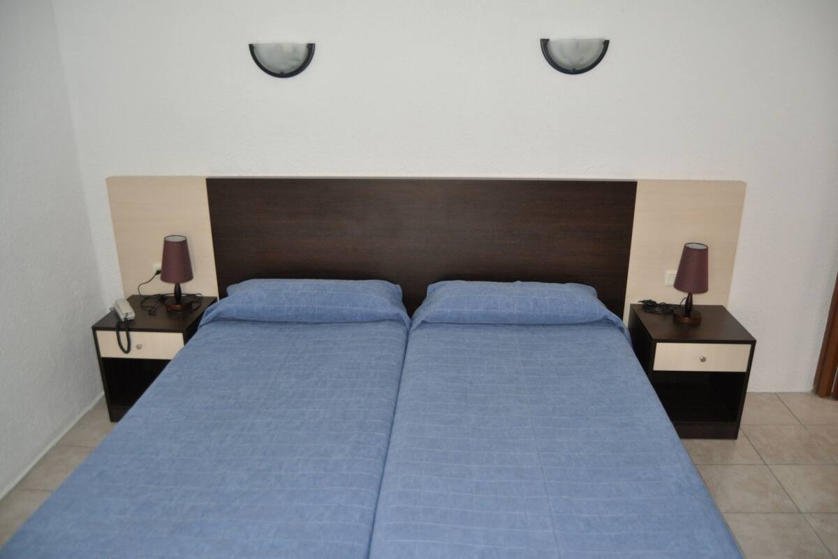 Una cama doble con cabecera de madera, dos mesitas de noche con lámparas y una pared blanca lisa.