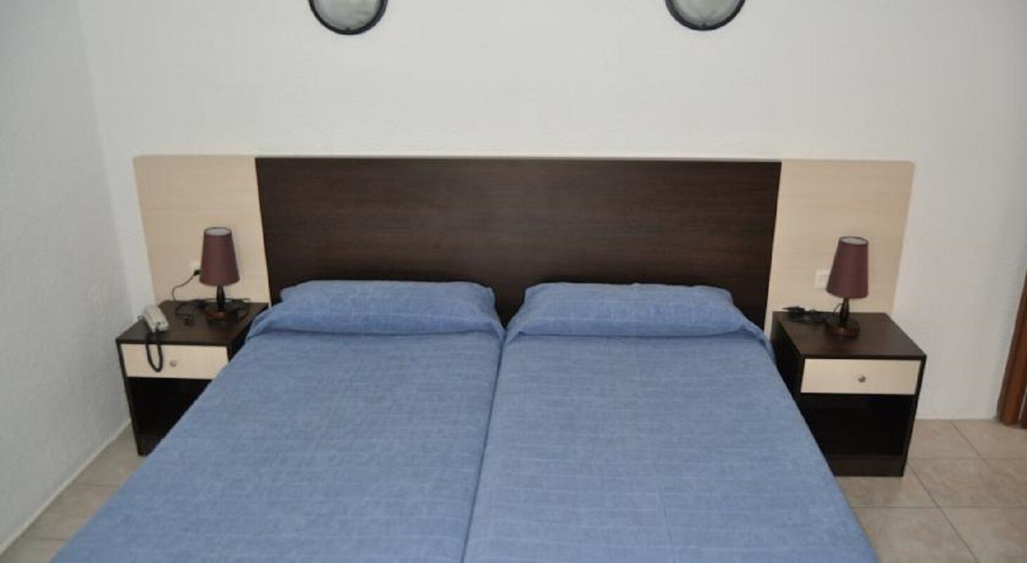 Una cama doble con cabecera oscura, dos mesitas de noche con lámparas y una pared lisa.