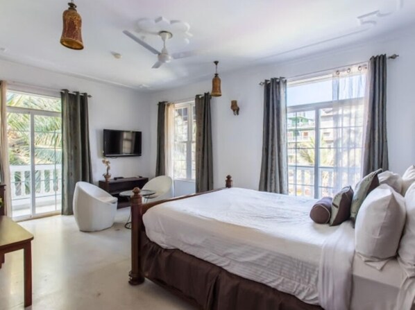 A bedroom with a large bed, a desk, a chair, a television, and a ceiling fan. - Safira River Front Resort (Morjim)