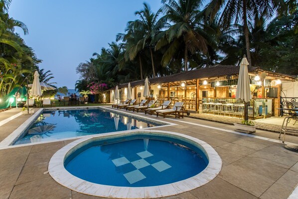 Una piscina con sillones de playa y sombrillas, rodeada de palmeras y un edificio con terraza.