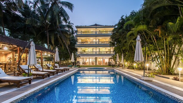 A swimming pool with lounge chairs and umbrellas, surrounded by palm trees and a multi-story building with balconies. - Safira River Front Resort (Morjim)